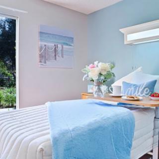 A cozy bedroom featuring a blue and white color scheme, with striped bedding and decorative pillows. A small table holds flowers, and natural light streams in through a nearby window.