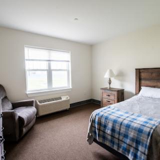 A simple and clean bedroom featuring a single bed, a nightstand with a lamp, a comfortable chair, and natural light coming through the window.