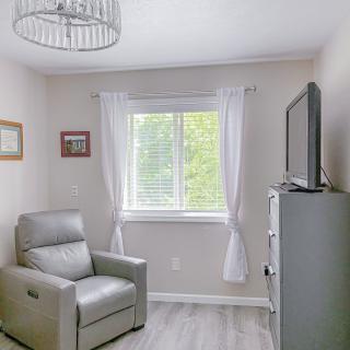A cozy minimalist living room with a grey armchair, TV stand, and a window with white curtains.