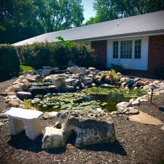 A serene garden pond surrounded by rocks in front of a house, with water lilies and a stone bench nearby.