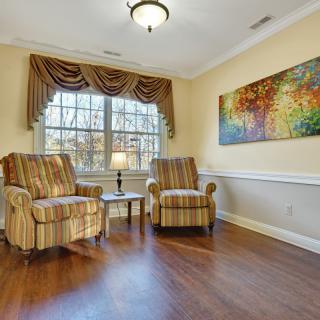 A cozy living room with two striped armchairs flanking a small table and lamp, set against a large window with draped curtains. Colorful artwork adorns the wall, and wooden floors add warmth to the space.