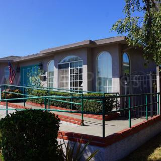 A single-story house with a ramp leading to the entrance, surrounded by a small garden under a clear blue sky.