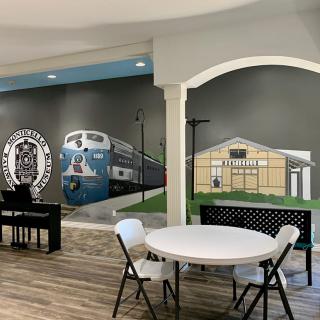A spacious room with a mural depicting a train and station, featuring a round dining table with chairs and a piano.