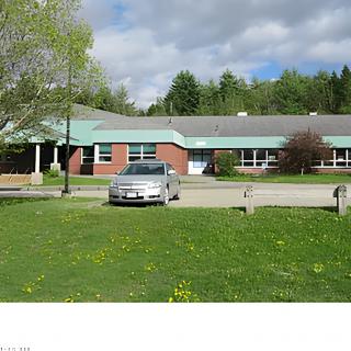 A school building with a car parked in front, surrounded by trees and grass under a cloudy sky.