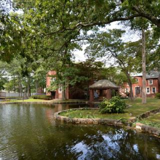 Scenic view of a pond with surrounding trees and a brick building in the background.