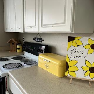 A bright and cheerful kitchen corner featuring a sunflower-themed artwork, a yellow breadbox, and an inviting cooking space.