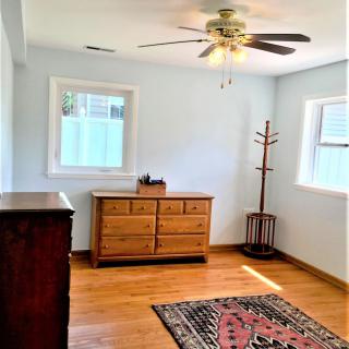 A bright bedroom with wooden floors, featuring a dresser, ceiling fan, and windows allowing natural light.