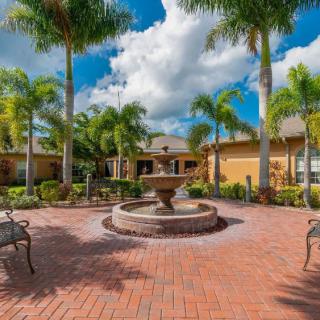 A tranquil courtyard with a central fountain, surrounded by palm trees and benches under a partly cloudy sky.