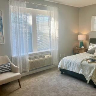 A cozy and modern bedroom with a neatly made bed, a comfortable chair, and natural light streaming in through a large window.