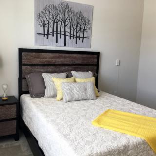 A modern bedroom with a stylish bed, featuring yellow and gray pillows, and a matching blanket. A piece of artwork with trees hangs above the headboard.