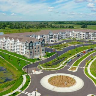 Aerial view of a modern apartment complex surrounded by lush greenery, featuring a roundabout and expansive open fields.