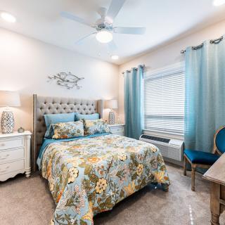 A cozy bedroom with elegant decor and furniture, featuring a bed with floral bedding, a nightstand with a lamp, and large windows with teal curtains.