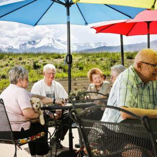 A group of seniors enjoying a gathering on a patio with colorful umbrellas and a scenic mountain view in the background.