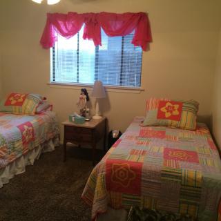 A cozy bedroom with two twin beds featuring colorful bedspreads, separated by a nightstand under a window with red curtains.