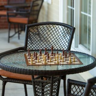 An outdoor chessboard set up on a glass-top patio table with chairs around it.