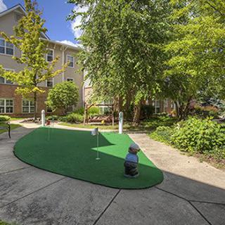 A sunny courtyard featuring a mini golf putting green surrounded by lush greenery and trees.