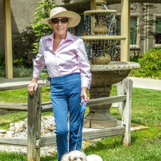 A woman in a hat and sunglasses stands by a garden fountain, accompanied by her dog.