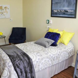 A cozy bedroom setup featuring a single bed with colorful pillows and a soft blanket, complemented by a chair and wall artwork.