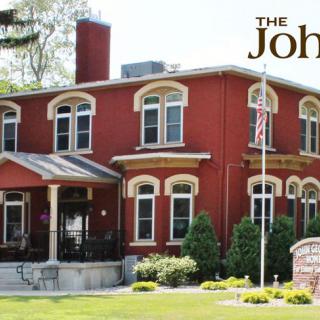 A historic brick building with arched windows and a flagpole in front, surrounded by trees and a manicured lawn.