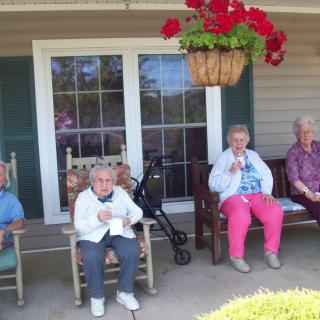 A group of elderly people sitting on a porch in rocking chairs, enjoying the outdoors under a hanging plant.