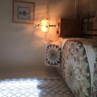 A cozy bedroom with a patterned quilt and pillows, illuminated by a wall-mounted lamp.