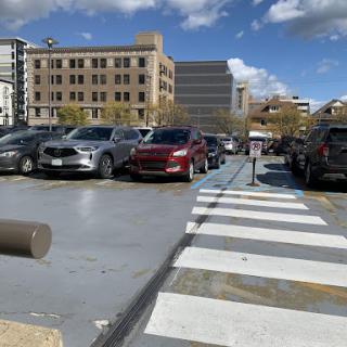 A bustling parking lot with various cars, set against a backdrop of urban buildings and a partly cloudy sky.