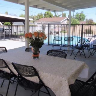 An outdoor patio with a table covered by a cloth, surrounded by chairs and decorated with a flower vase, overlooking a fenced swimming pool in the backyard.