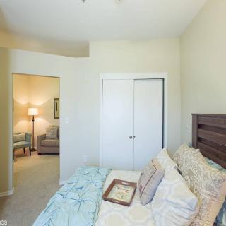 A cozy bedroom featuring a neatly made bed with pillows, a nearby closet, and an adjoining room with a lamp.