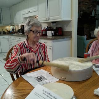 An elderly woman playing a drum in a kitchen setting.