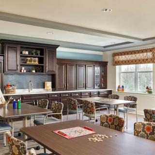 A spacious and well-lit kitchen area with multiple tables and chairs, featuring wooden cabinets and a large window.