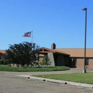 A one-story building with an American flag flying outside, surrounded by trees and a lawn under a clear blue sky.