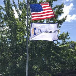 An American flag and a Diversicare flag flying on a flagpole against a backdrop of trees and a blue sky.