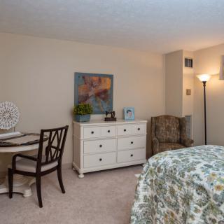 A cozy bedroom featuring a chair, lamp, dresser, and table with decor.