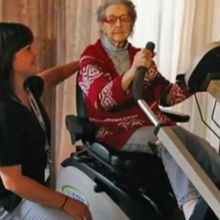An elderly woman using exercise equipment with the assistance of a caregiver.