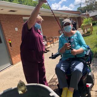 A caregiver assisting a person in a wheelchair with fishing on a sunny day.