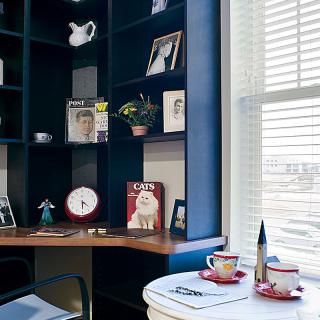 A cozy office corner with shelves displaying magazines and decor, a table set for coffee, and large windows letting in natural light.