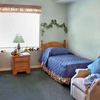 A cozy bedroom with a single bed, nightstand, lamp, and chair next to a window.