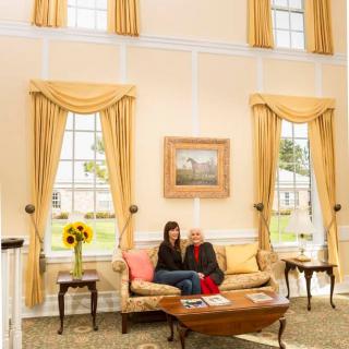 A cozy living room with two people sitting on a couch, surrounded by large windows with yellow curtains and elegant traditional decor.