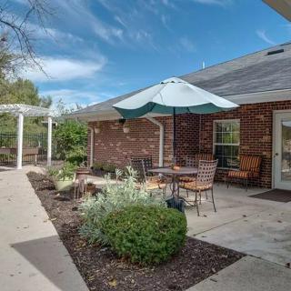A cozy patio area with outdoor seating and an umbrella, adjacent to a brick building surrounded by well-maintained landscaping.