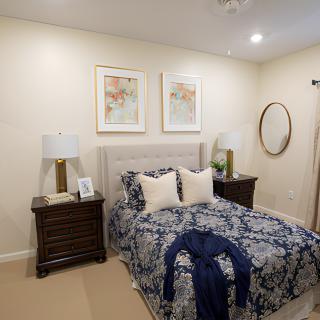 A cozy and elegantly decorated bedroom featuring a bed with patterned bedding, nightstands with lamps, framed art on the wall, and a round mirror.