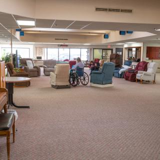 A spacious lounge area with various chairs and a wheelchair on a carpeted floor, illuminated by large windows.