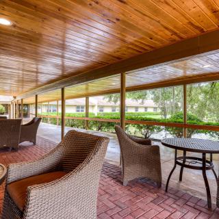 A cozy outdoor patio area with wicker furniture and tables, set on a brick floor with a wooden ceiling, overlooking a green landscape.