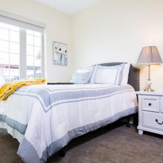 A cozy bedroom with a neatly made bed, featuring a bedside lamp and nightstand next to a large window.