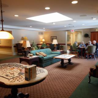 A cozy living room with comfortable seating and a table, featuring people gathered in conversation under warm lighting.