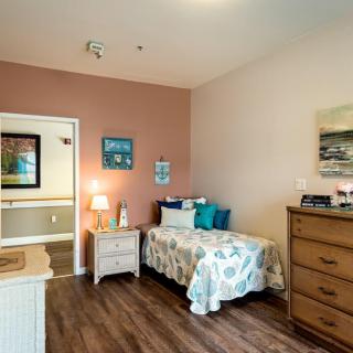 A cozy bedroom featuring a single bed with floral bedding, a nightstand with a lamp, and a wooden dresser on hardwood floors.