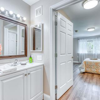 A bright and modern bathroom with a vanity mirror, leading into a bedroom.