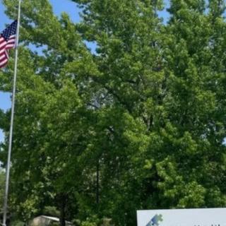 An American flag flies next to a sign surrounded by lush green trees under a clear blue sky.