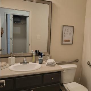 A tidy bathroom with a large mirror above the sink, neatly arranged toiletries, and a small piece of wall art.