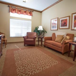 A cozy living room with a sofa, armchair, and a decorative rug, featuring framed artwork and a large window.