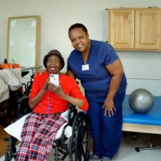 An elderly woman in a wheelchair smiling alongside a caregiver in a healthcare setting.